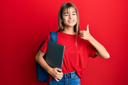 Teenager caucasian girl wearing student backpack and holding computer laptop smiling happy and positive, thumb up doing excellent and approval sign
