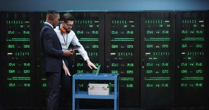 Young technician carries shelf with laptop showing necessary servers to his African-American boss in suit. Two IT workers check serviceability of computer servers.