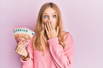Beautiful young caucasian girl holding bunch of 10 euro banknotes covering mouth with hand, shocked...