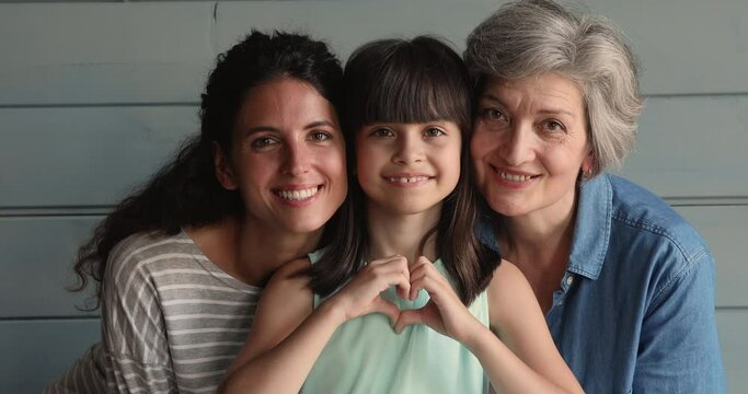 Little cute girl showing heart shape with fingers while pose in studio with elderly grandmother and mother smile look at camera. Three gen family portrait, life value, attachment and bonding concept