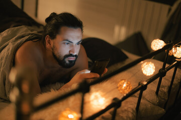 Obraz premium Young man with phone in bed in the evening. Evening leisure at home