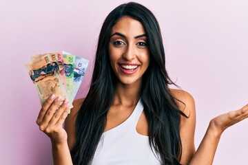 Beautiful hispanic woman holding canadian dollars celebrating achievement with happy smile and winner expression with raised hand