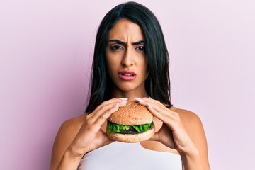 Beautiful hispanic woman eating a tasty classic burger clueless and confused expression. doubt...