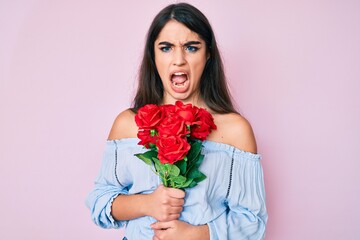 Brunette teenager girl holding flowers in shock face, looking skeptical and sarcastic, surprised with open mouth