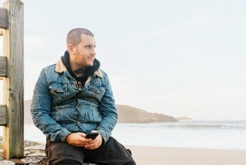 man with tattoo and piercings poses with a serene gesture in front of the beach and holding his mobile phone.