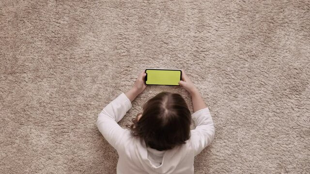 kid girl using phone technology device lying on carpet floor alone. Small child hold pad computer surfing internet play game at home. Children tech addiction concept