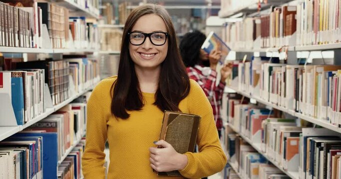 Portrait of young happy female nerd student standing in library holding book and smiling looking at camera.
