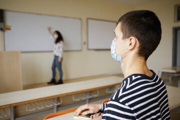 Young male student wearing a face mask and and listening to female teacher in classroom. New normal. Social distancing in schools and universities due to the Coronavirus pandemic. Safety measures.