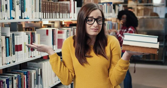 Close up portrait of young female student with glasses standing in library and frustrated looking at stack of books to read for lecture.