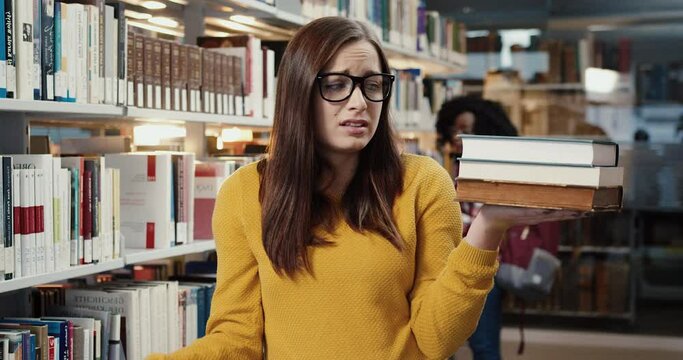 Portrait of unfortunate student stands in library and fearfully examines the number of books that need to be read quickly.