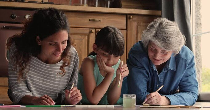 Three gen family diverse women granny mom and child spend weekend together gathered in kitchen lying on warm floor hold paint brushes drawing on paper enjoy leisure. Kid development home hobby concept