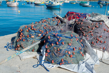 Fishing nets with red floats and blue ropes rest on a dock under bright sunlight, with colourful...