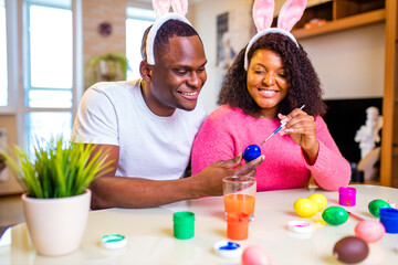 Young african american man and woman wearing cute easter bunny ears and painting eggs in the house