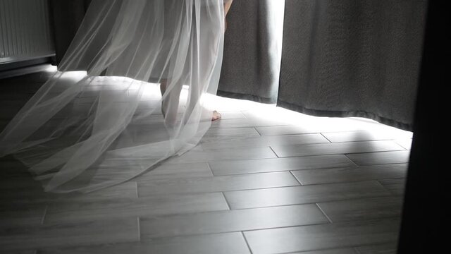 Close-up of the sexy legs of a young girl step on the parquet floor. The bride with a robe and a long veil comes to the window and opens the curtains.