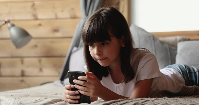 Adorable little girl lying on bed in cozy bedroom holding smartphone, spend free weekend time on internet, watch on-line vlog channels, younger generation addicted with gadgets, device overuse concept