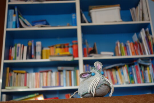 Selbstgebastelte Stoffspielzeug Maus vor buntem chaotischen  B&uuml;cherregal. Lesemaus mit blauen Augen, Schnurrbarthaaren und treuem Hundeblick. Library Laboratory rat or mouse science concept image .