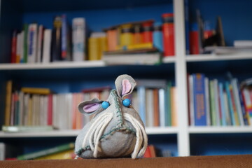 Slebstgebastelte Stoffspielzeug Maus vor buntem chaotischen  Bücherregal. Lesemaus mit blauen Augen, Schnurrbarthaaren und treuem Hundeblick.