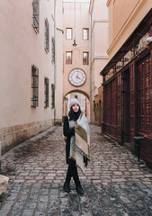 A young girl in coat, angora hat, wide scarf and autumn boots is walking along a narrow old street with paving stones waiting for a date. Lviv, Armenian courtyard, Ukraine.