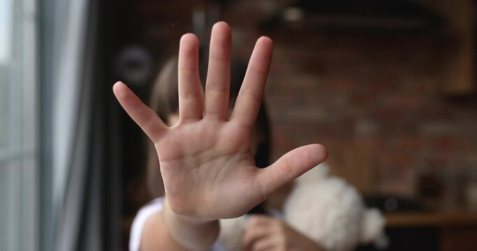 Little lonely girl standing indoor holds stuffed toy outstretching palm to camera makes protective gesture. Social issues, stop child abuse, bullying, discrimination, violence against children concept