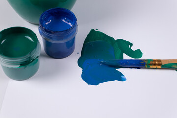 two open jars of gouache next to each other, and on a white surface next to a few strokes of green and blue, and a dirty brush lies on them, next to a jar of water