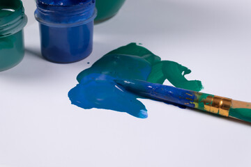 a stained brush lies on strokes of green and blue paint, and next to it are open jars of these same colors