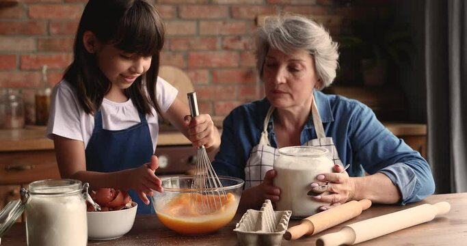 60s elderly female babysitter teach little girl to cook. Loving grandmother and small granddaughter prepare family recipe cake for event, stirring eggs make dough enjoy process. Hobby, cookery concept