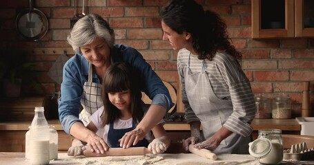 Three gen women in apron cooking together in kitchen, elderly granny, grown up daughter little granddaughter prepare pie, flattening dough enjoy process. Transfer skills from generation to gen concept