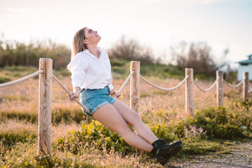 chica joven rubia con pantalones cortos vaqueros azules y camisa blanca sentada en unas cuerdas mirando al cielo