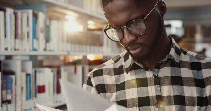 Close up of young smart African American student standing in library and intently reading book in preparation for exams.