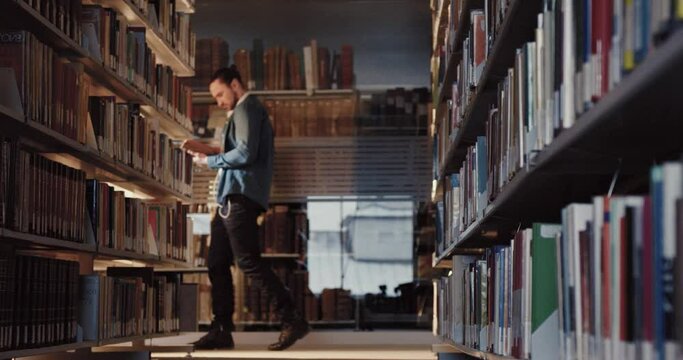 University library in evening time. Young male student walks among the bookshelves intently reading book. Distance learning.