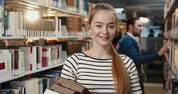 Close up portrait of young blonde female student standing in library holding books and looking at camera smiling.