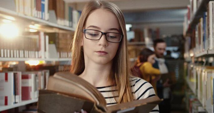 Close up of young beautiful caucasian blonde female student standing in library pulls book off the shelf and starts reading.