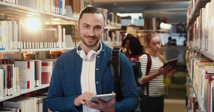 Portrait of young successful Caucasian student standing in the library among bookshelves looking through the list of necessary literature on tablet looks up and smiles at camera.