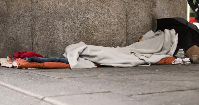 Sleeping Place Of A Homeless Man On The Urban Street With Blankets, Umbrella, Clothes, And Food - Slider Right, Low-Level Shot