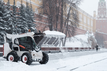 A small loader excavator bobcat removes snow from the sidewalk near the Kremlin walls during a...
