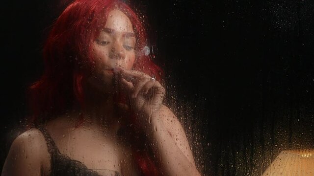 Sexy multi-ethnic woman in red wig smokes marijuana cigar dressed in black lace lingerie looking through a rain soaked window