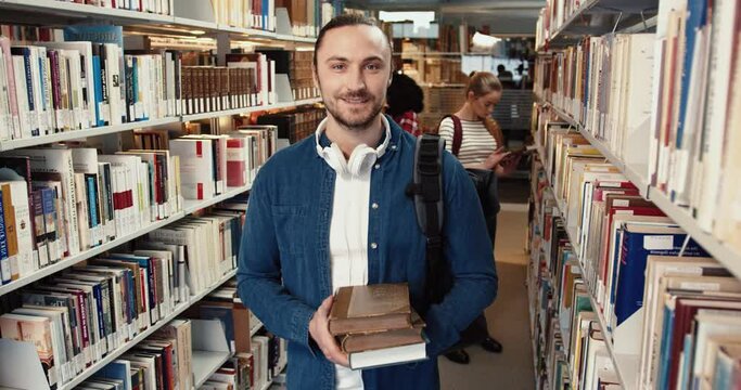 Portrait of young stylish guy in denim shirt standing in library holding stack of books and smiling at camera.