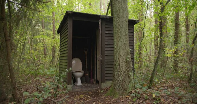 Shed with a toilet in the middle of the woods. Outside lavatory. Outside toilet in the forest. Old Shack with toilet bowl and gear. Primitive bathroom with open door in an outbuilding between trees.