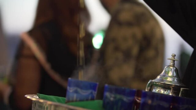 Close up shot of Tea Pouring Into A Glass With Moroccan Tea Set after sunset outdoors. Blurred background. Slow motion shot.
