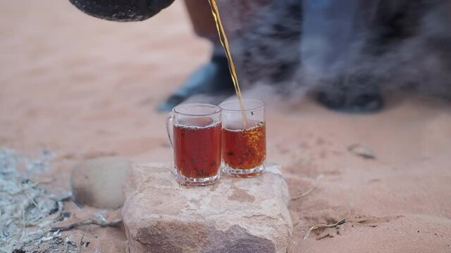 A bedouin pouring hot tea in some glasses, while camping in the desert near a rudimentary campfire.
