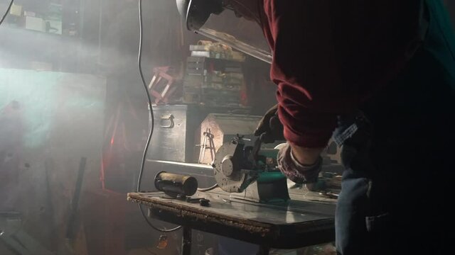 A worker sharpens a tool on a bench grinder in a smoke-filled workshop. Sparks fly from the grinding wheel.
