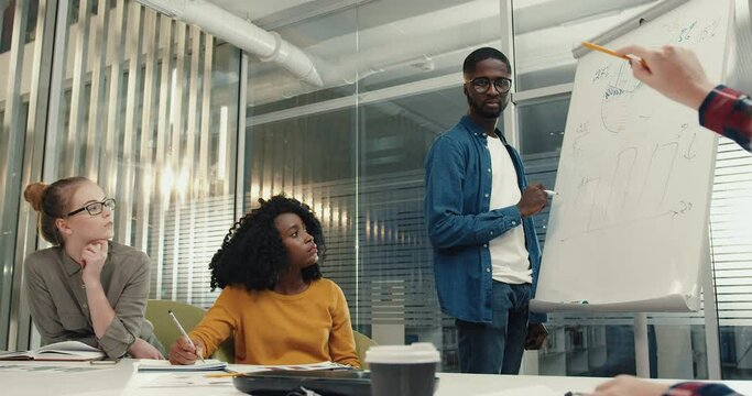 Students sit in classroom and discuss solution of problem. Young African-American guy with glasses stands at board and enters data.