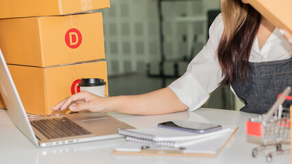 A young Asian female startup owner looks happily looking at her laptop with a box at home preparing for package delivery in the sme concept, omnichannel purchasing.