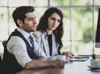 Side view portrait of a thoughtful young adult caucasian businessman sitting in a meeting, thinking how to solve company problems. Selective focus at an absent-minded man with blurred businesswoman - obrazy, fototapety, plakaty