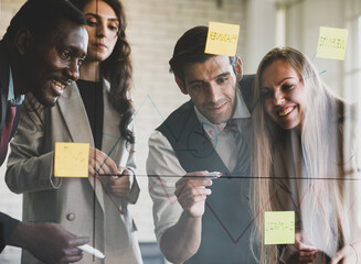 Group of four professional multiracial businessmen and businesswomen brainstorming and discussing by writing graph with a marker on glass wall to explain company financial status to plan strategy
