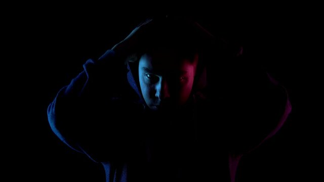 A young man takes off his hood. An attacker is standing in the dark. Blue and red light falls on the person on a black background.