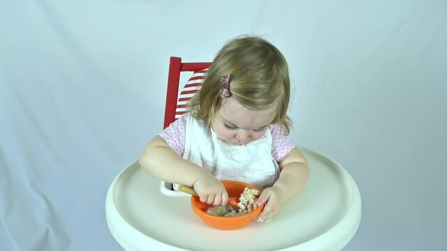 An adorable caucasian female toddler takes a big bite of oatmeal porridge with a wooden spoon from a plastic bowl. She is sitting on a high chair and wearing a bib