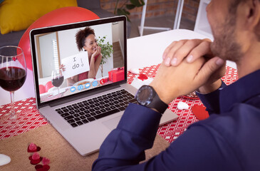 Diverse couple making valentine's date video call the woman on laptop screen holding i do sign