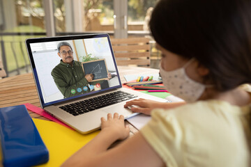 Fototapeta na wymiar Caucasian schoolgirl wearing face mask using laptop on video call with male teacher