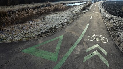 Bycicle road near river bank with frozen grass visible, winter season, morning sunshine. Symbols of bike and triangular right of way painted green on asphalt are visible. 